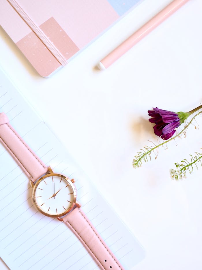 services-01 Flat lay of a minimalist pink watch with a flower and notebook on a white background.